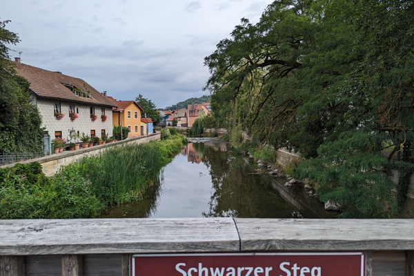 Das Bild zeigt den Blick von der Brücke "Schwarzer Steg" auf den Fluss Haßlach. Es ist ein Hinweisschild angebracht. Im Hintergrund sieht man Häuser der Stadt.