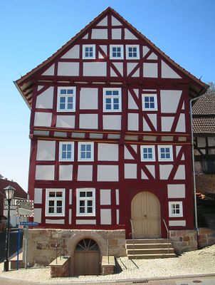 Außenansicht eines viergeschossigen Fachwerkhauses mit rot-braunen Balken und großer, abgerundeter Eingangstür aus hellem Holz. Der Sockel ist aus Sanstein, ein Kellerabgang führt zu einer großen Tür mit halbrundem Glaseinsatz als oberem Teil.