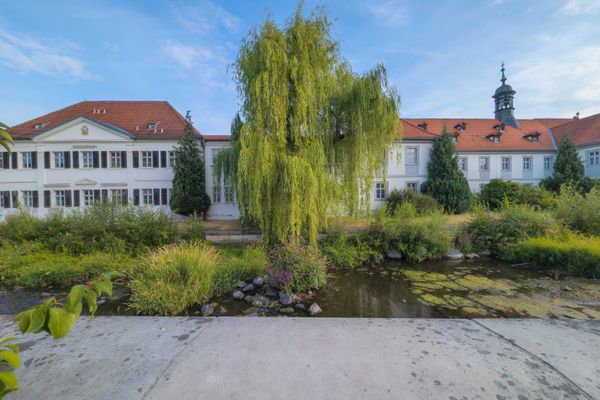 Das Bild zeigt die Spitalkriche St. Anna in Kronach. Im Vordergrund ist ein Fluss zu sehen.