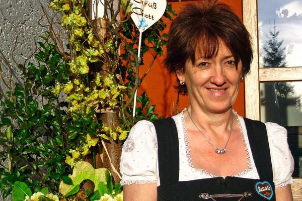 Die Gastgeberin, bekleidet mit einem Dirndl, steht freundlich lächelnd vor dem Eingangsbereich. Hinter ihr stehen auf einem Schild in Herzform die Worte "Grüß Gott".