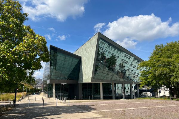 Modernes Gebäude mit schräg geneigter Glasfassade, umgeben von Bäumen und gepflastertem Platz unter blauem Himmel mit Wolken.