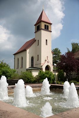 Wild aufschäumend plätschern einige Fontainen des Brunnens, der mit Sandsteinen rechtekig gefasst ist. Dahinter ist leicht unscharf die kleine, weiße Ev. Kirche mit ihrem roten Spitzdach zu sehen. Sie ist umgeben von grünen und roten Laubbäumen und ragt in den blauen, leicht bewölkten Himmel hinein.