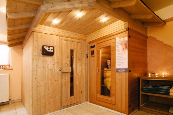 Im Haus ein originelles, mit einem Dach versehenes Saunagebäude aus Holz. In der Infrarotkabine sitzt eine, mit einem Handtuch bekleidete Frau. Teelichter auf der Handtuchkommode verströmen ein angenehmes Licht.