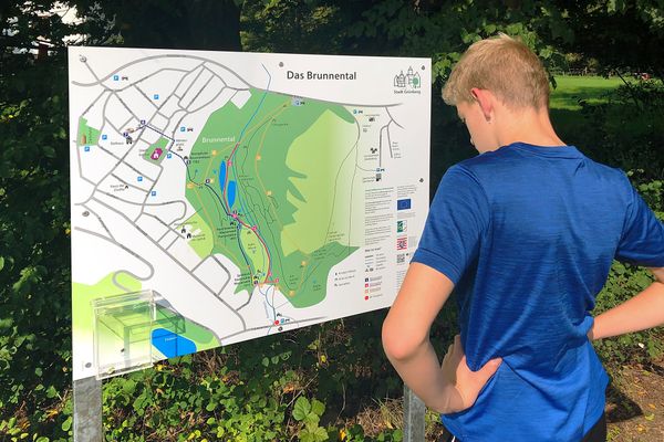 Ein junger Mann steht in Freizeitkleidung vor einem großen Infoschild mit der Wegeführung durch das Brunnental.