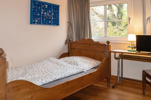 Einzelbett mit Bettwäsche neben einem Fenster, daneben ein Schreibtisch mit Lampe und Fernseher sowie ein Stuhl.