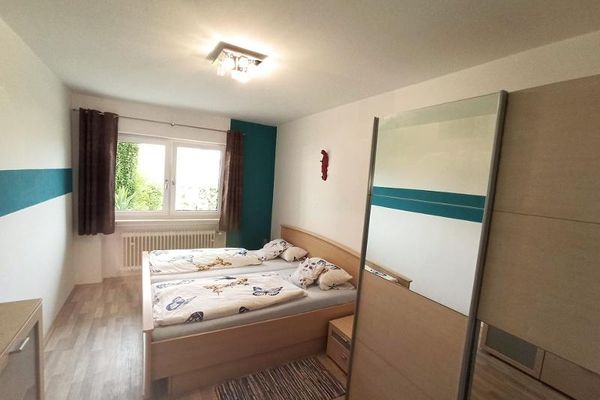 Ein Holz-Doppelbett mit Nachttisch steht im Raum, dazu ein Schiebetürenschrank und eine Kommode. Helle Wände mit grünen Akzenten. Am Doppelfenster hängen dunkle Vorhänge, vor dem Bett liegt ein kleiner Flickenteppich.