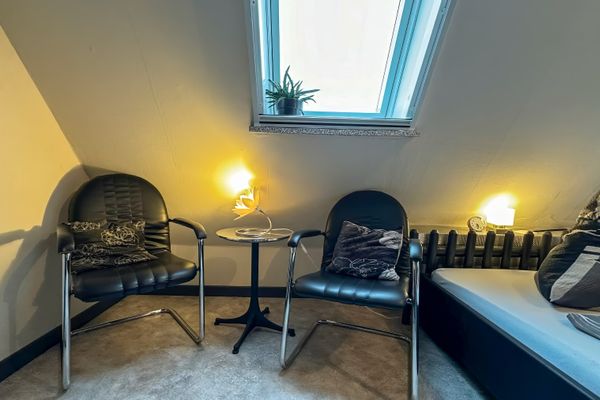 Zwei schwarze Sessel mit Metallgestell stehen unter einer weißen Dachschräge. Dazwischen befindet sich ein kleiner, runder Tisch mit einer eingeschalteten Lampe in Blütenform. Über der Sitzgruppe ist ein Dachfenster zu sehen. Rechts im Bild ist der Rand eines Bettes sowie ein Heizkörper erkennbar.