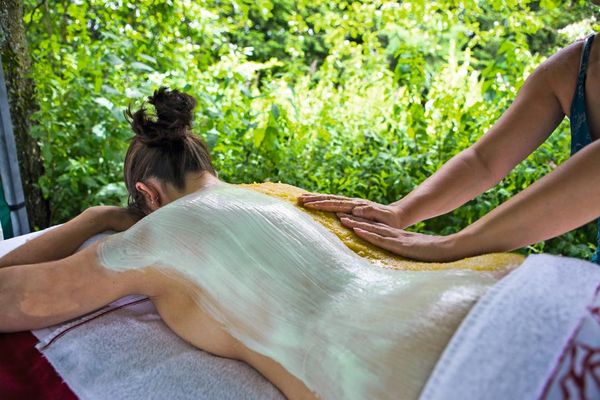 Eine Dame liegt auf einer Massagebank, dahinter wachsen dichte, grüne Pflanzen. Eine Rückenhälfte ist dick eingecremt, die andere Hälfte wird mit Peelingmaterial massiert.