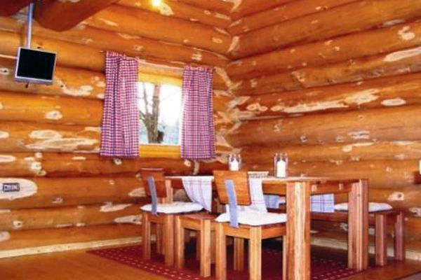 Vor einer Innenwand aus Naturstammholz steht auf einem rot gemusterten Teppich ein massiver Holztisch mit Bank und Stühlen, vor einem Fenster mit rot karierten Vorhängen. An der Wand ein Flachbildfernseher.