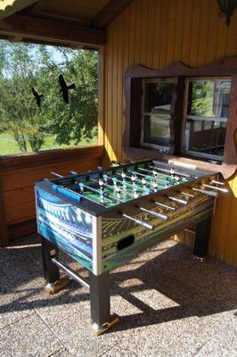 Vor dem Überdach eines Holzgebäudes steht ein Kickerkasten. Das doppelte Sprossenfenster ist mit einem geschwungenen Holzrahmen verziert.