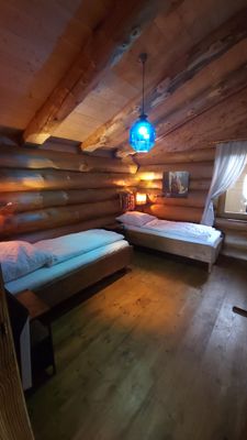 Schlafzimmer im Holzhaus mit zwei L-förmig gestellten Einzelbetten. Eine blaue Deckenlampe hängt über dem Raum, helle Vorhänge rahmen das Fenster.