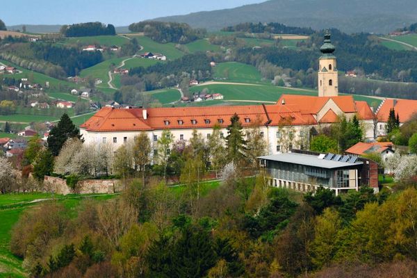 Aus einiger Entfernung bietet sich ein Blick auf die beeindruckende Klosteranlage mit Klosterkirche in Windberg. Hohe Bäume stehen vor dem Gästehaus. In der hügeligen Landschaft im Hintergrund liegen zwischen Wiesen und Waldflächen verstreut mehrere Gebäude.