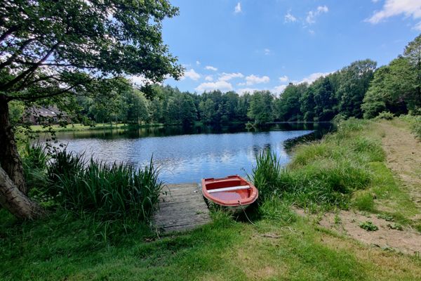 Dorfteich in Räber (Suderburg) mit spiegelnder Wasseroberfläche, umgeben von Bäumen; im Vordergrund Holzsteg mit rotem Ruderboot, Grasufer und schmaler Sandweg rechts, blauer Himmel mit wenigen Wolken.