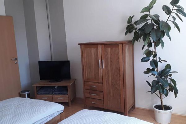 Nebeneinander stehen ein TV-Tisch mit Fernseher, eine Holzkommode und eine Grünpflanze. Im Vordergrund sind die Bettenden zweier Einzelbetten zu sehen.