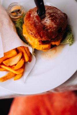 Auf dem Bild ist ein Burger und Pommes auf einem Teller zu sehen. Ein Messer steckt in dem Burger (zur Befestigung). Daneben steht eine kleines Glas mit Sauce und einem Löffel.