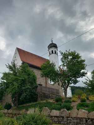 Eine kleine Kirche mit eckigem Turm und Zwiebelkuppel steht erhöht und teils verdeckt von hohen Sträuchern. Davor eine gepflegte Fläche mit niedrigen Pflanzen, eingerahmt von einer markanten Steinmauer.