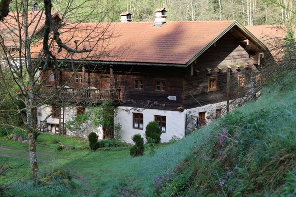 Hinter einem alten Bauernhaus mit zwei Kaminen, gemauertem Untergeschoss und hölzernem Obergeschoss erhebt sich eine grasbewachsene Böschung. Das Haus und zwei Nebengebäude sind von Laubbäumen umgeben.