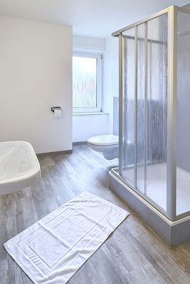 Das Bild zeigt ein Badezimmer mit einer Dusche auf der rechten Seite, die durch eine halbtransparente Duschkabine abgetrennt ist. Links ist ein wandmontiertes Waschbecken zu sehen, und im Hintergrund befindet sich ein WC. Die Wände sind weiß, der Boden mit Fliesen in Holzoptik ausgelegt, und ein Fenster mit Milchglas sorgt für Tageslicht.