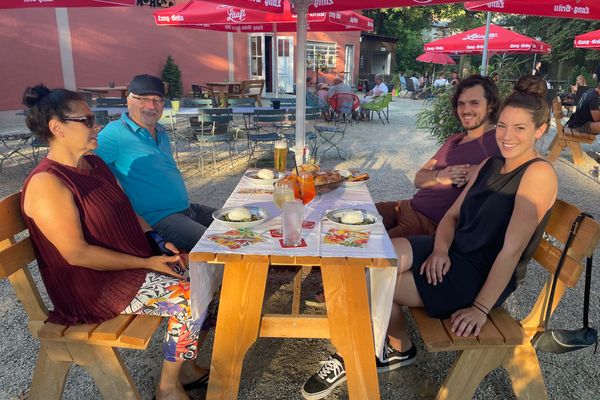 Auf dem Bild sind vier Personen an einem Tisch auf Bänken sitzend. In der Mitte des Tischs befindet sich ein Sonnenschirm. Auf dem Tisch stehen Getränke und Brotzeiten.
