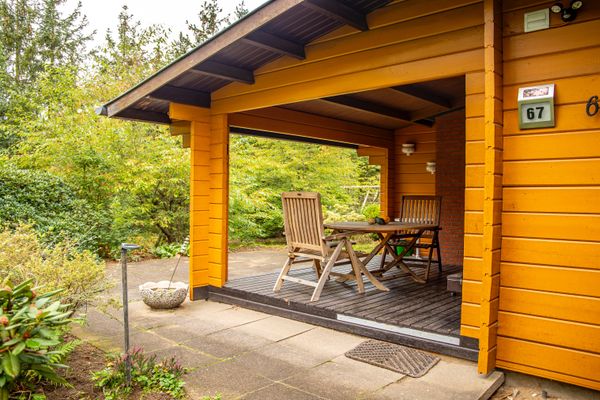 Überdachte Terrasse an einem Holzhaus mit Tisch und Stühlen, angrenzend an gepflasterten Außenbereich und Garten.