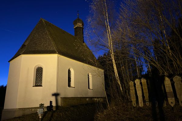 Die Leonhardikapelle unter tiefblauem Himmel in der Abenddämmerung. Daneben stehen vor hohen Bäumen mehrere aufgereihte Totenbretter.