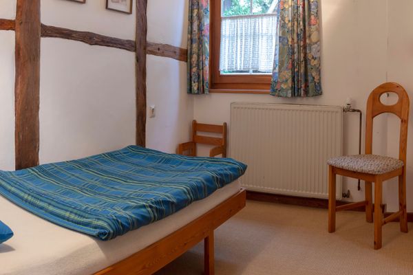 Schlafzimmer mit Einzelbett, Stuhl, Fenster mit Vorhang und Heizkörper unter dem Fenster.