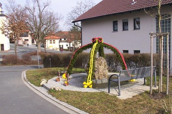 Der Osterbrunnen mit roten und gelben Eiern steht zwischen der Straße und einem Haus.