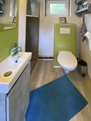Badezimmer mit Waschbecken links, WC rechts und Dusche im Hintergrund, Fenster darüber.