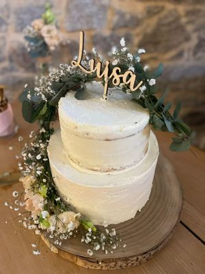 Auf dem Bild ist eine weiße, 2 stöckige Torte zu sehen. Auf der Torte steckt ein Namensschild aus Holz (Luisa). An der Seite ist die Torte mit einer Blumengirlande verziert (grün, weiß).