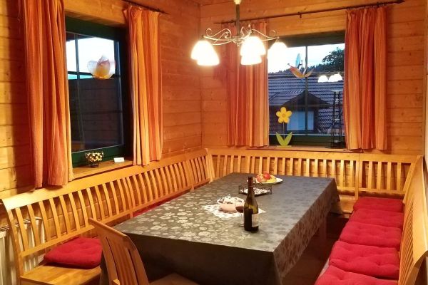 U-förmige Holzeckbank mit Sprossenrückwand, Tisch und Stuhl. An der holvertäfelten Wand befinden sich zwei Fenster, durch die man die Nachbargebäude sehen kann.  Die Vorhänge sind  rotgemustert.