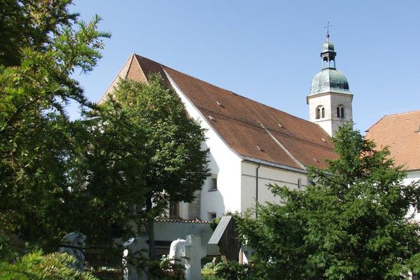 Unter blauem Himmel im Vordergrund Grabsteine unter hohen Bäumen, dahinter ein Teil der Wallfahrtskirche Bogenberg mit Kirchturm.