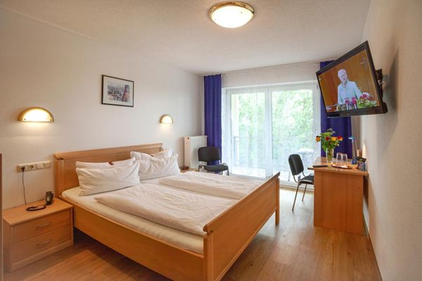 Ein Schlafzimmer mit Doppelbett im Holzrahmen, flankiert von Nachttischen. Über dem Bett hängt ein Bild zwischen zwei Wandlampen. Im Raum stehen ein Schreibtisch mit Blumenvase und zwei Gläsern sowie zwei Stühle. Ein TV-Gerät ist an der Wand befestigt. Vor der Doppelbalkontüre hängen blaue Vorhänge.