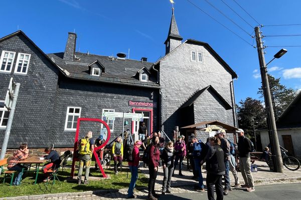Im Hintergrund ein großes Haus mit Schieferfassade. Im Vordergrund steht eine Gruppe Wanderer
