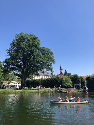 Eine großzügige Parkanlage mit Teich und Schloss ist zu sehen. Es ist sommerlich warm und die Menschen flanieren. Auf dem Teich sind Kanufahrer unterwegs. Im Hintergrund ist ein roter Kirchturm zu sehen.