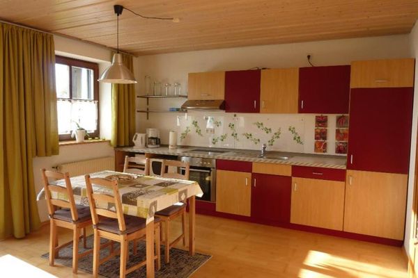 Bunte Küchenzeile mit versetzt angebrachten holzfarbenen und roten Türen sowie Kaffeemaschine, Wasserkocher und Wandablageregal mit Dekogläsern. Davor ein Holztisch mit Tischdecke und vier Stühle. Über dem Tisch hängt eine runde, graue Lampe, am Fenster grüne Vorhänge.