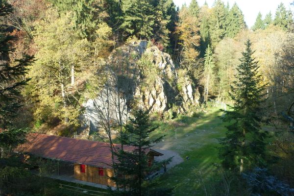 In einer Senke, umgeben von einer sonnendurchfluteten Felsformation und Mischwald, liegt ein langgezogenes Holzgebäude mit Ziegeldach.