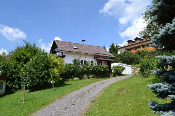 Zwischen einer Rasenfläche führt eine kiesbedeckte Auffahrt zu einem von Sträuchern teils verdeckten Haus. Im Hintergrund ein weiteres Gebäude mit Dachgauben.
