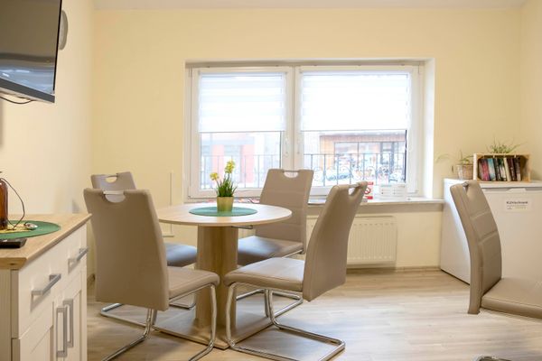 Raum mit rundem Holztisch und vier hellen Stühlen vor einem Fenster, seitlich Kühlschrank mit Bücherregal und Dekoration.