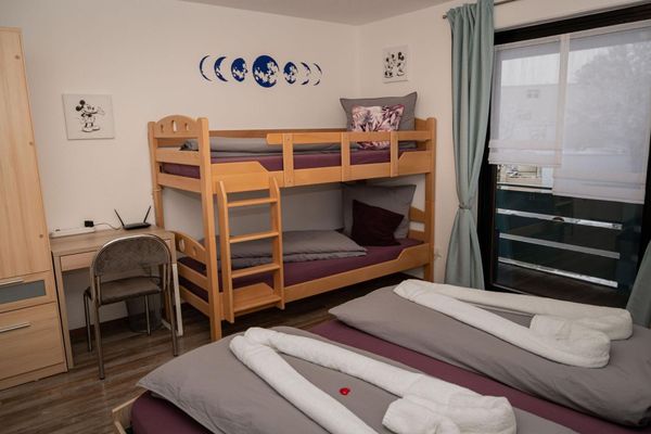 Im Schlafzimmer stehen ein Doppel- und ein Stockbett, ein kleiner Schreibtisch und ein Schrank. Die Betten sind lila und grau bezogen und schön dekoriert. Bilder und Dekomalerei schmücken die Wand. An der Fensterfront befinden sich helle Raffrollos und lindgrüne, bodenlange Vorhänge.