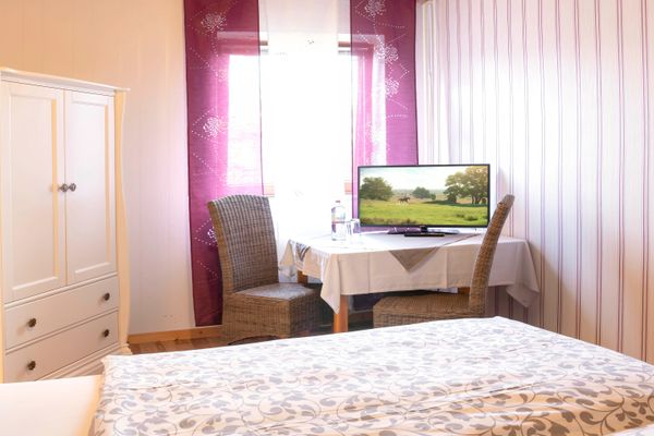 Blick in ein Gästezimmer auf eine Sitzecke vor einem Fenster. Ein Tisch mit weißer Tischdecke und einem darauf stehenden Flachbildfernseher wird von zwei braunen Korbstühlen flankiert. Links steht ein weißer Kleiderschrank mit zwei Schubladen. Im Vordergrund ist ein Bett mit gemusterter Bettwäsche angeschnitten. Das Fenster ist mit bordeauxroten Vorhängen dekoriert.