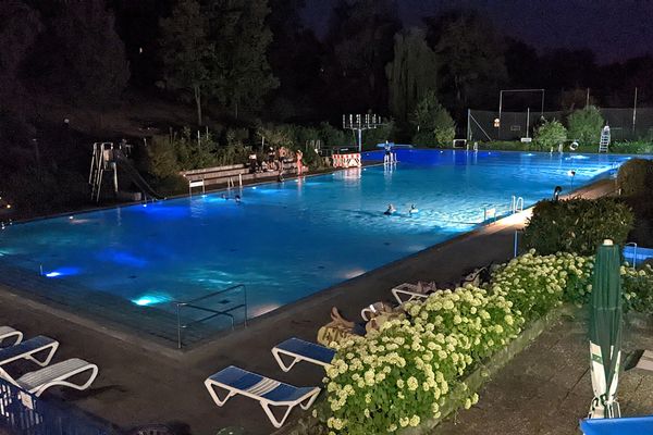 Es ist Nacht. Das rechteckige Becken des Schwimmbades ist vom Wasser aus beleuchtet, so dass das Wasser blau strahlt. Vier Menschen sind im Wasser, ein paar stehen am Beckenrand. Hinter dem Becken stehen Bäume und am vorderen Rand stehen auf großen Gewegplatten weiße Liegestühle. Eine Hecke aus fast zartgrünen Hortensien trennt den Beckenbereich  von einer Terrasse ab, auf der die Sonnenschirme zugeklappt sind.