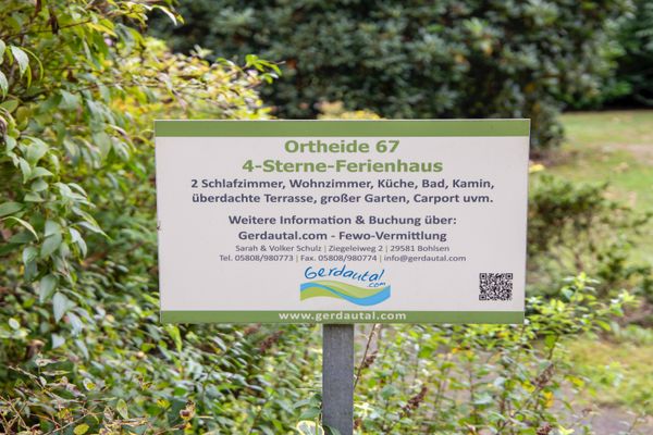 Informationsschild mit Text zu einem Ferienhaus, aufgestellt vor Sträuchern und Rasenfläche.