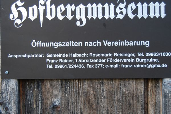 An einer verwitterten Bretterwand hängt eine Schild mit der Aufschrift "Hofbergmuseum". Das Schild gibt Auskunft über Öffnungszeiten und Ansprechpartner.
