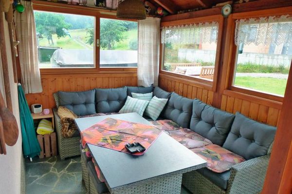 Im holzvertäfelten Wintergarten stehen ein graues Rattan-Ecksofa mit Tisch und ein Beistelltischchen. An den Fenstern hängen Bistrogardinen und helle Vorhänge. Über den Fenstern sind Dekorationsartikel aufgereiht.