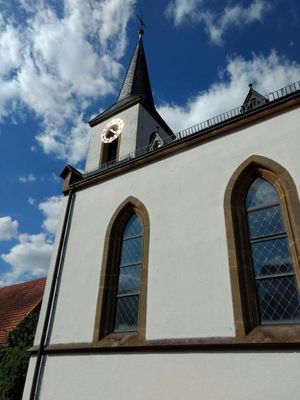 Die goldene Kirchturmuhr glünzt im Sonnenlicht. Der blaue, leicht bewölkte Himmel spiegelt sich in den großen Fenstern.