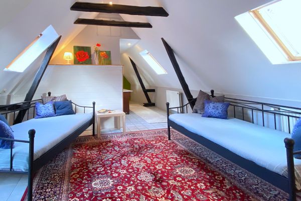 Ein helles Zimmer im Dachgeschoss mit weißen Wänden, Dachschrägen und dunklen sichtbaren Holzbalken. Im Raum stehen sich zwei schwarze Metallbetten mit hellblauen Bezügen und blauen Kissen gegenüber, die auf einem großen, rot gemusterten Teppich platziert sind. Durch mehrere Dachfenster fällt Tageslicht in den Raum.