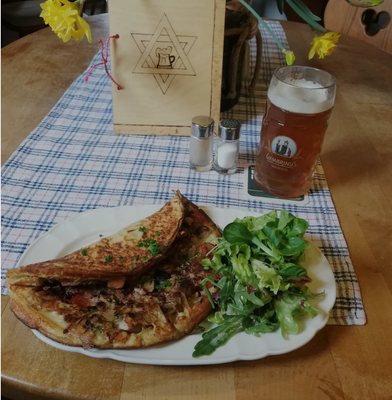 Auf dem Bild ist auf einem Tisch stehend ein Teller mit einem Omelett und einem Salat zu sehen. Dahinter steht auf einer Tischdecke ein Salz- und ein Pfefferstreuer. Daneben ist ein Glas Bier zu sehen. Im Hintergrund sind Blumen und eine Holztafel mit einem Brauerstern darauf zu sehen.