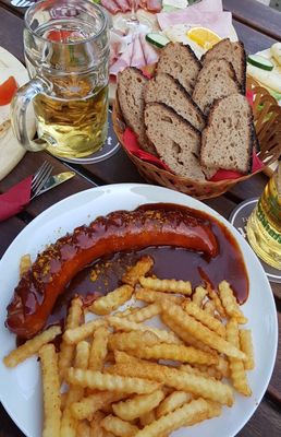 Auf dem Bild ist ein Gericht (Currywurst mit Pommes) zu sehen. Im Hintergrund befinden sich verschiedene Brotzeiten.