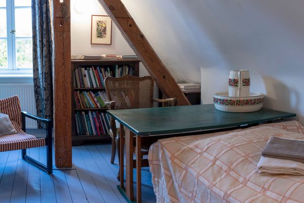 Bett mit kariertem Überwurf neben einem Tisch mit Tablett, dahinter ein Bücherregal und ein Stuhl.