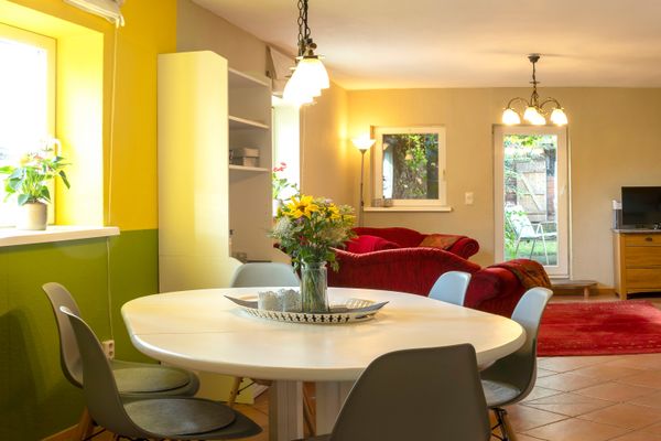 Runder Esstisch mit Stühlen im Vordergrund, dahinter Wohnzimmer mit Sofa und Fenster.